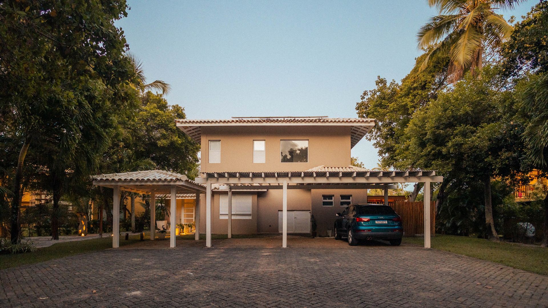 Exterior view of a modern house with a driveway and palm trees at sunset, offering a serene tropical ambiance.